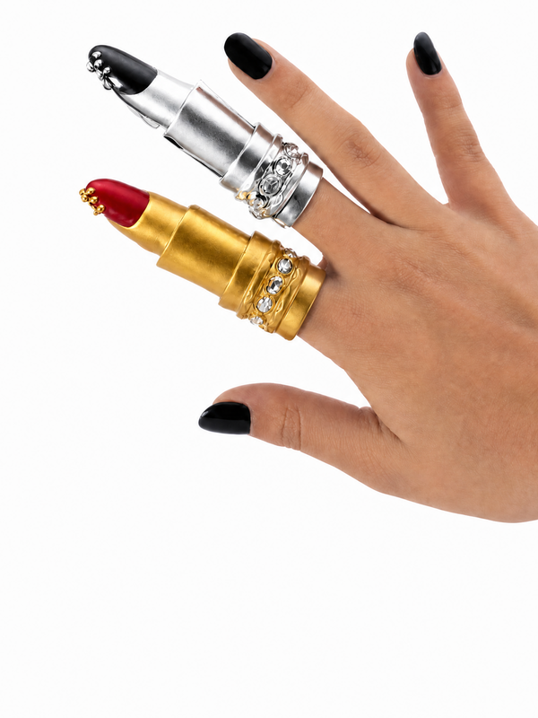 Gold and silver lipstick-shaped ring on a hand with black nail polish against a white background
