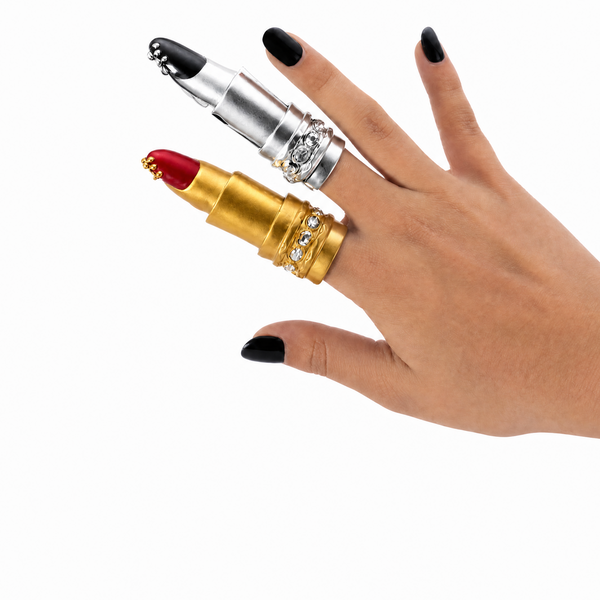 Gold and silver lipstick-shaped ring on a hand with black nail polish against a white background